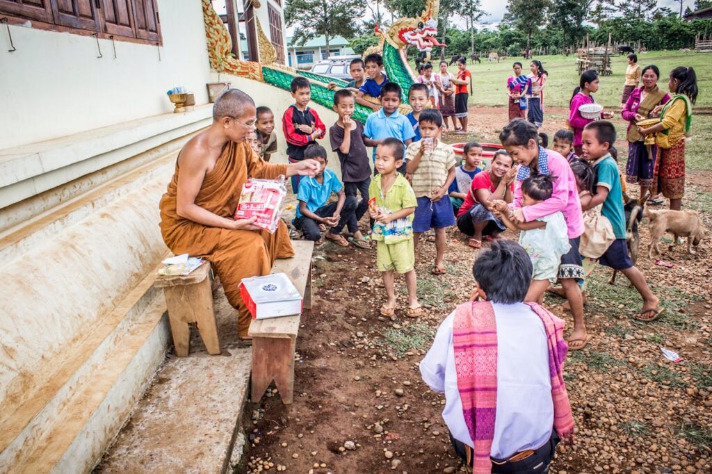 A Buddhist monk distributing items to a group of children and adults at a temple in a rural area.